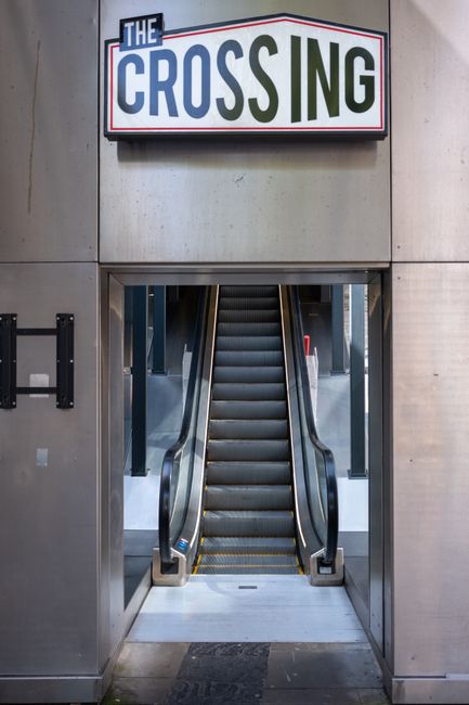 View of a staircase descending from a grey, unfinished ceiling with visible metal fixtures and electrical conduits, leading down to a tiled corridor at Guildford Station. The corridor features a yello