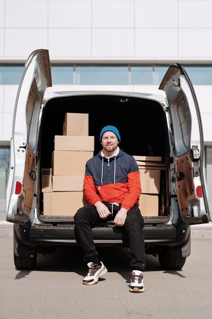 A man wearing a white T-shirt, jeans, and a black cap is standing on a residential street, loading a large cardboard box into the side door of a white commercial van with black accents, parked paralle